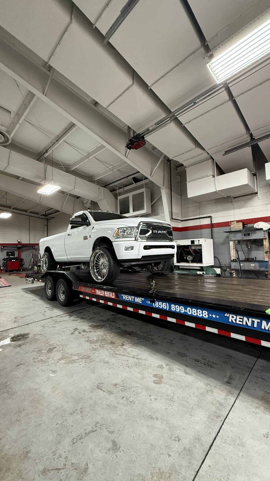 A white pickup truck sits on a flatbed trailer inside a spacious, brightly lit auto repair workshop.