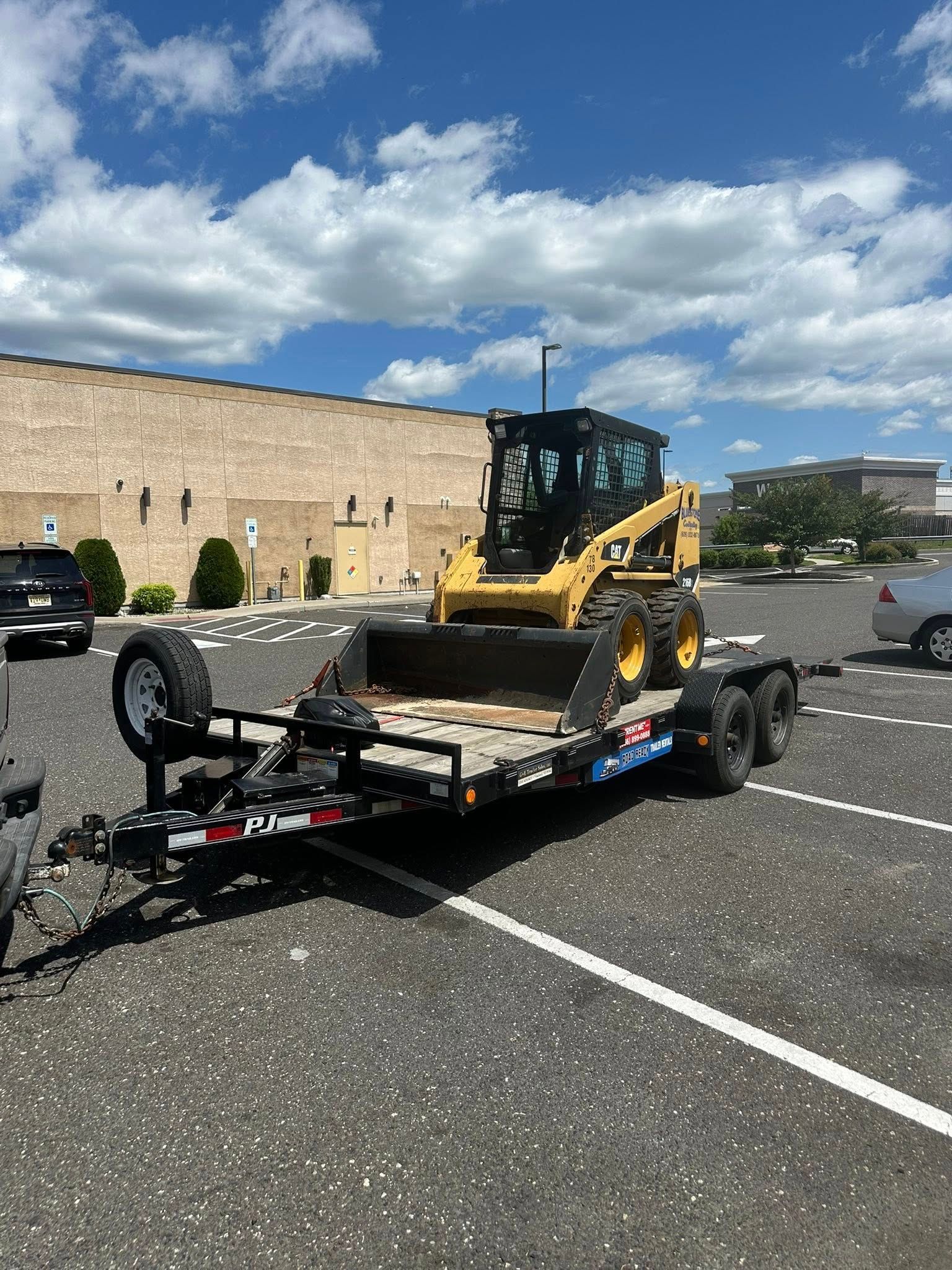 A yellow skid-steer loader loaded on a flatbed trailer parked in an asphalt parking lot under a blue, cloudy sky.