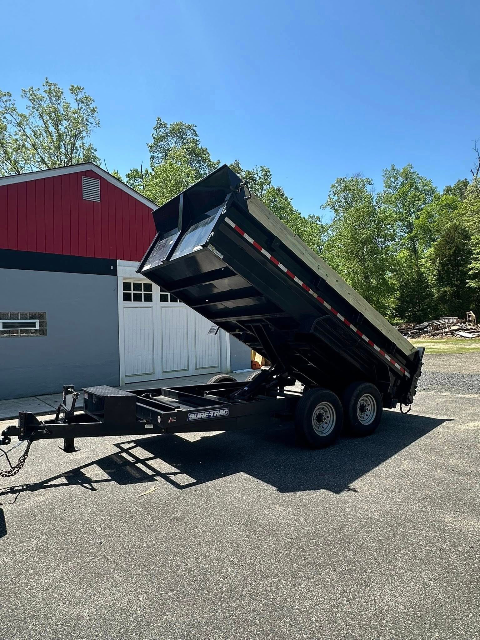 A black dump trailer with its bed raised, parked on a gravel lot next to a building with red and grey exterior walls.