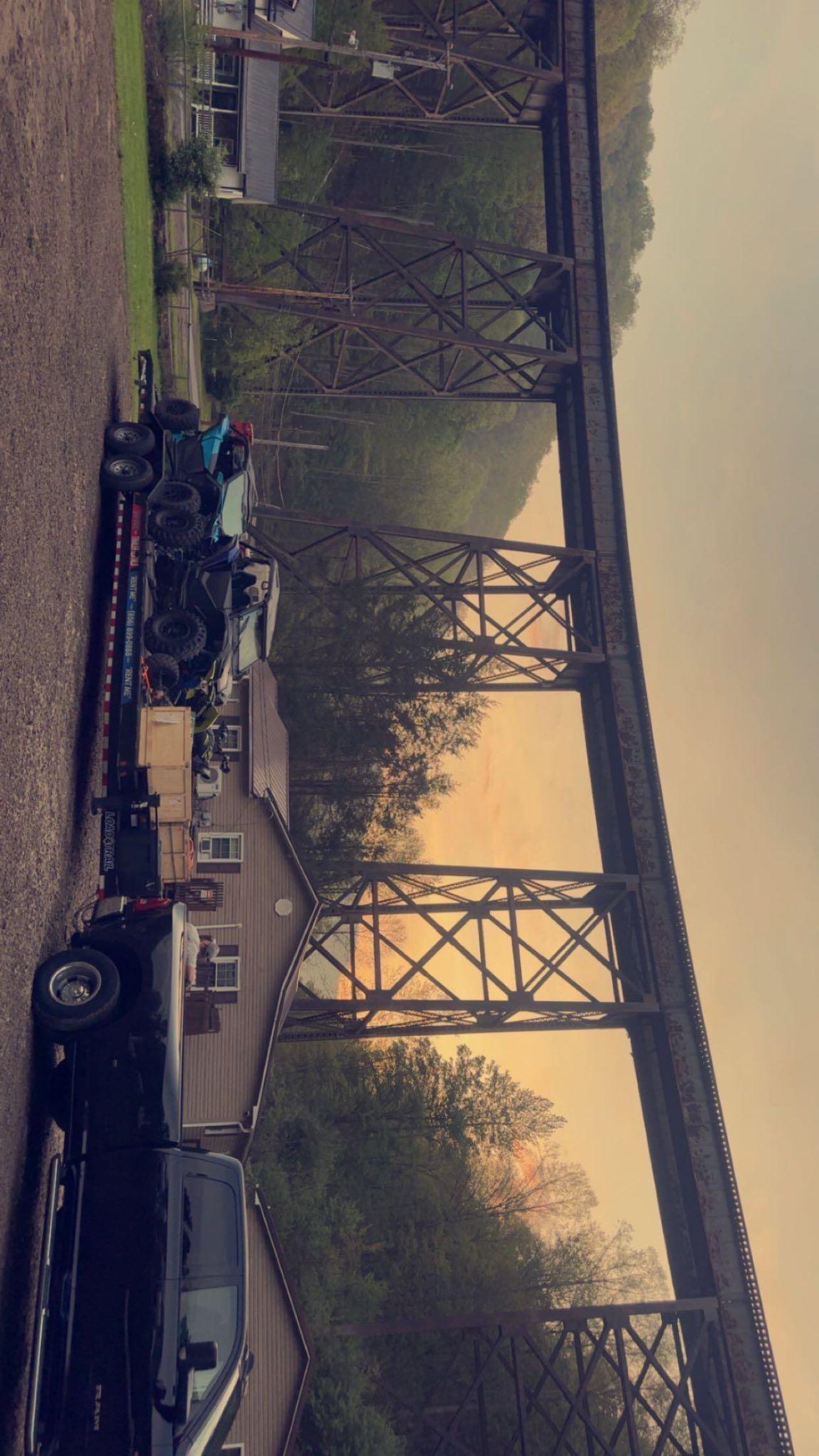 A large, dark steel railroad trestle spans over a rural setting at sunset, with a truck and trailer parked below.