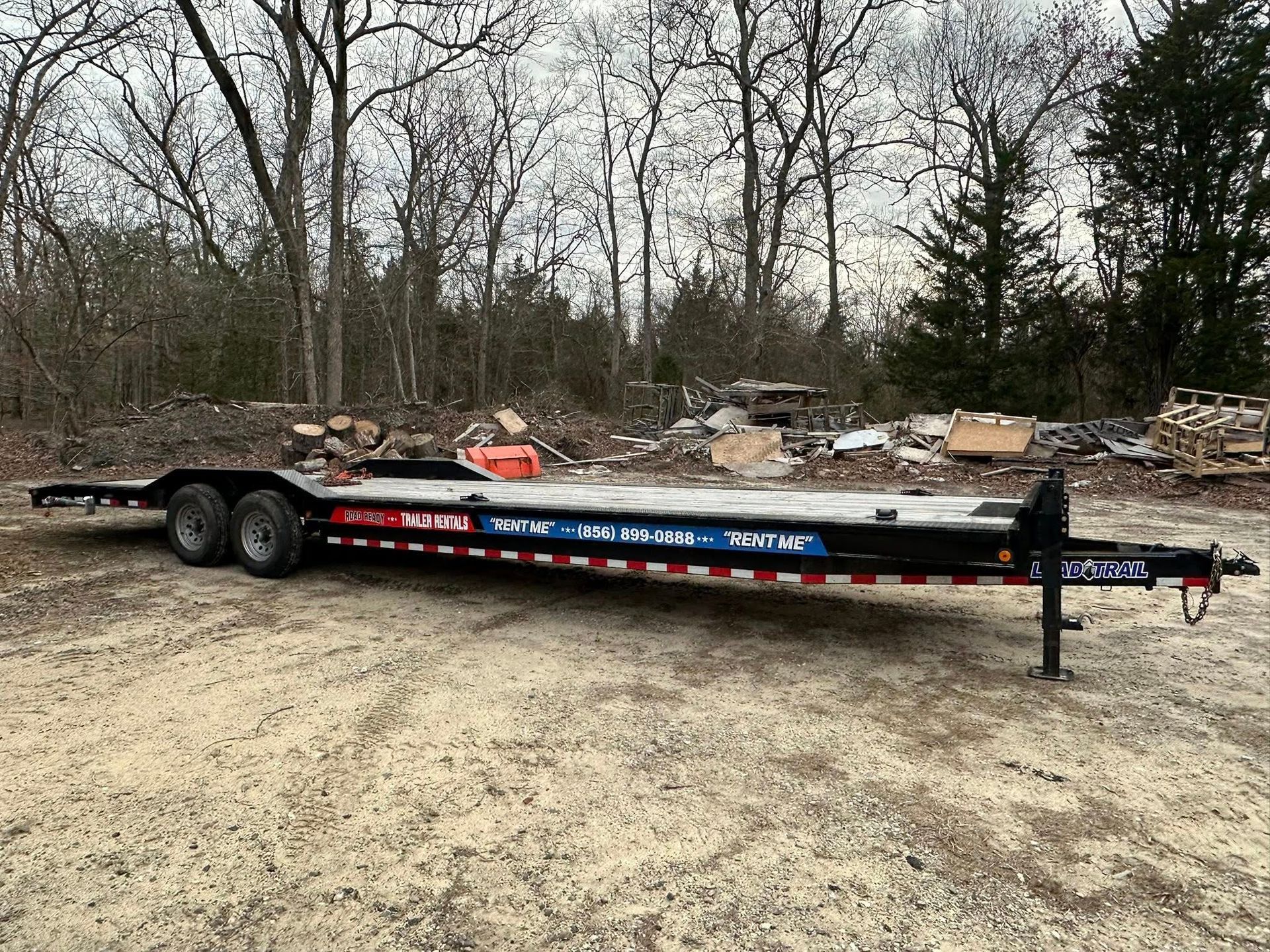 A black flatbed car hauler trailer parked on a gravel lot with trees in the background.