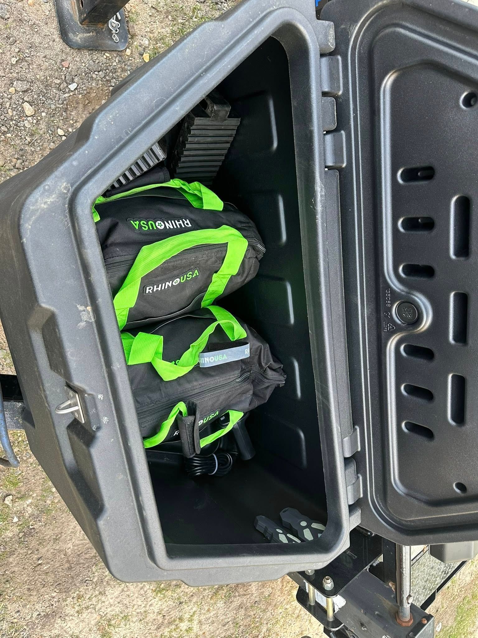 Open black storage box on a trailer tongue containing two black bags with neon green straps and a set of wheel chocks.