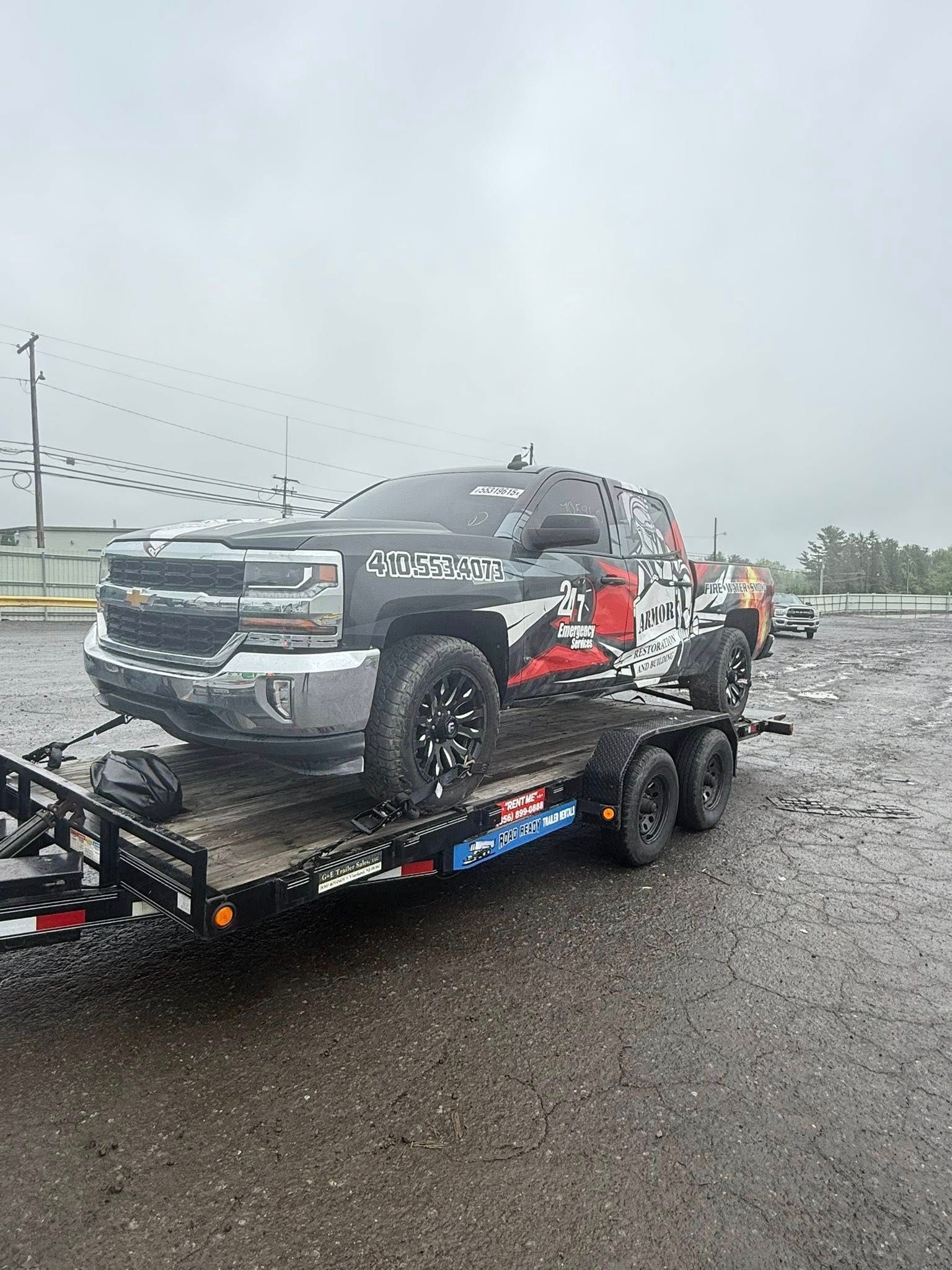 A black and white Chevrolet pickup truck with red decal accents on a flatbed trailer in a muddy, overcast lot.