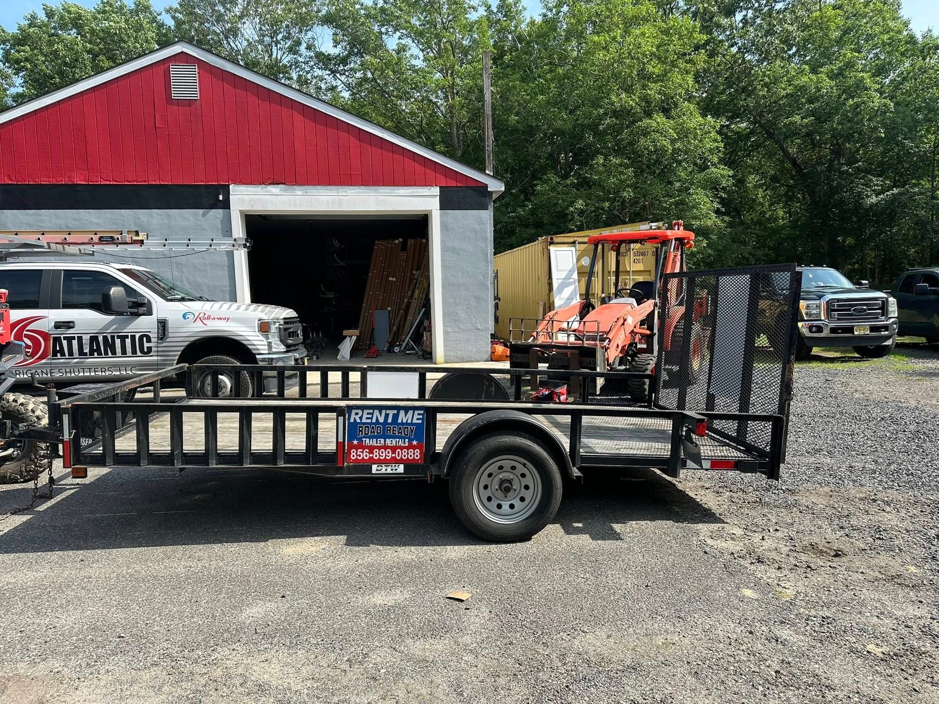 A black utility trailer parked on a gravel lot in front of a building with a red roof, next to a white pickup truck.