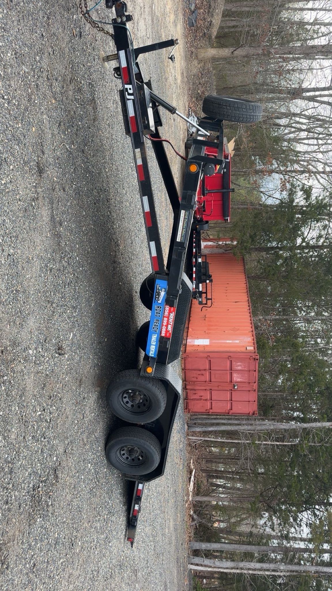 A black dual-axle equipment trailer parked on a gravel lot near a large orange storage container and a wooded area.