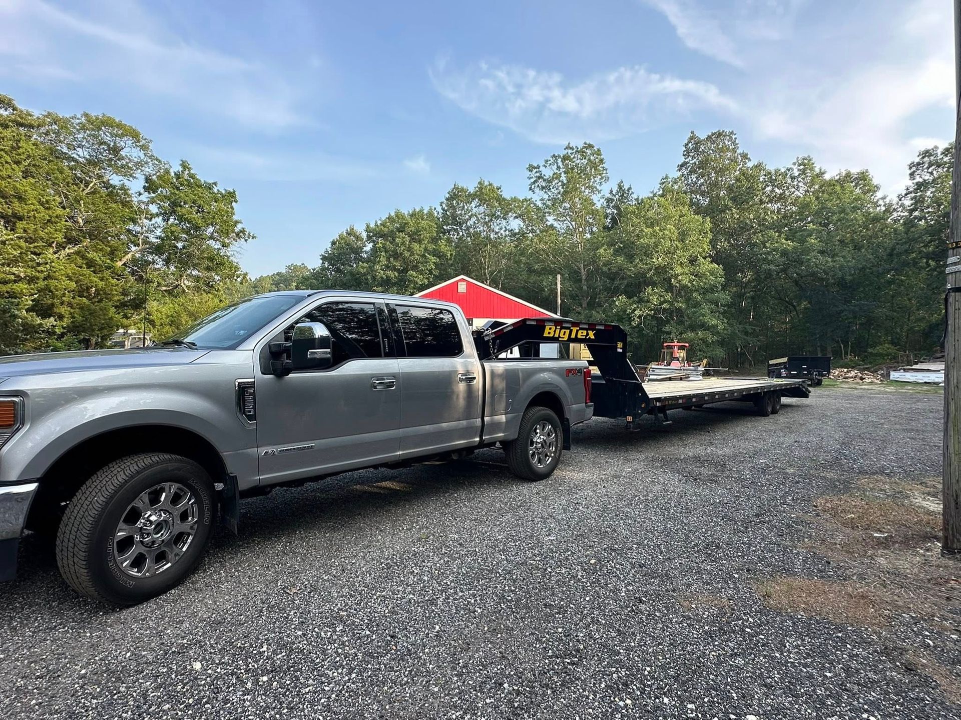 A silver pickup truck parked on a gravel lot, hitched to a flatbed gooseneck trailer under a clear blue sky.