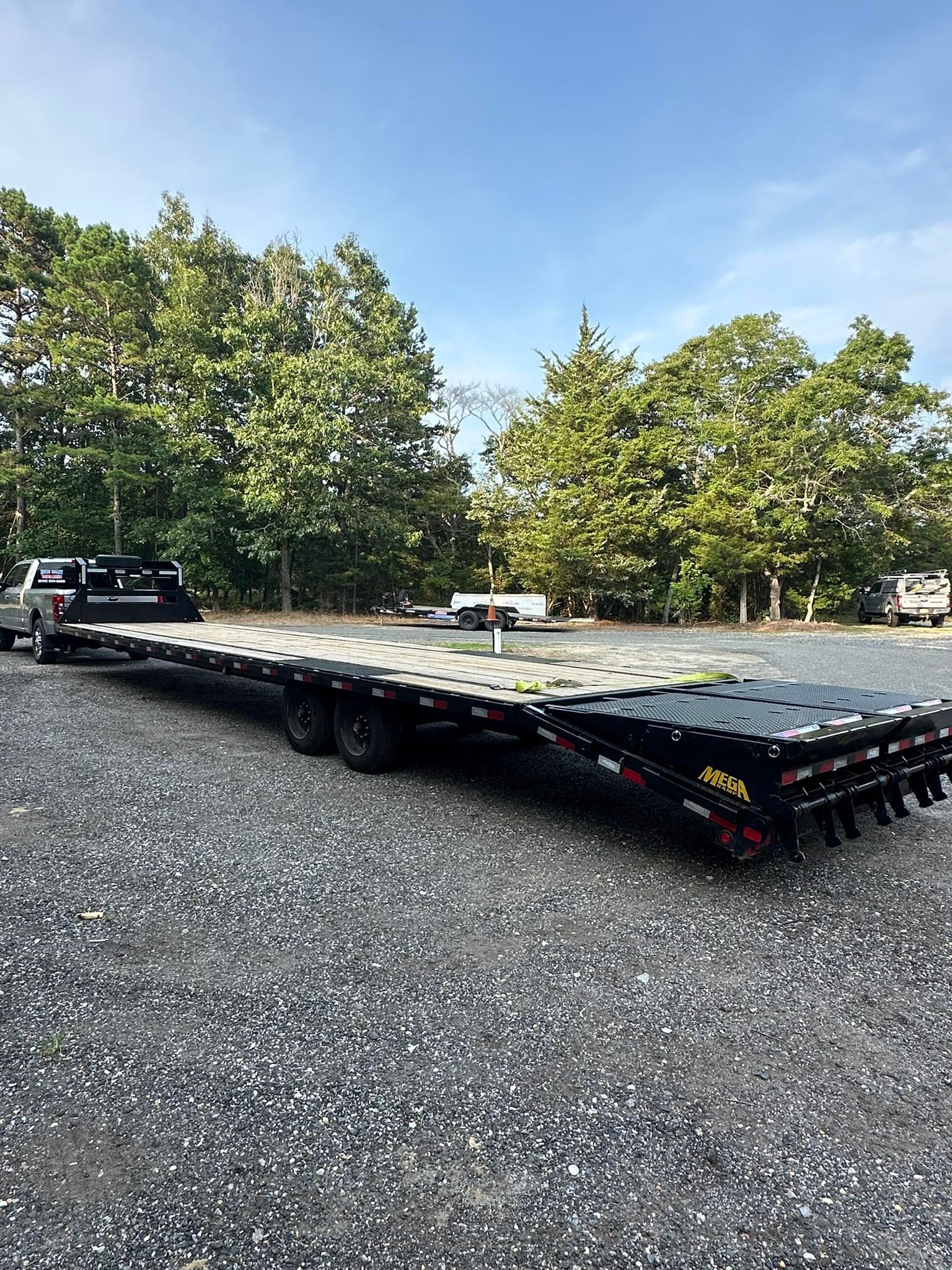 A black pickup truck towing a long, black flatbed trailer parked on a gravel lot near trees under a clear blue sky.