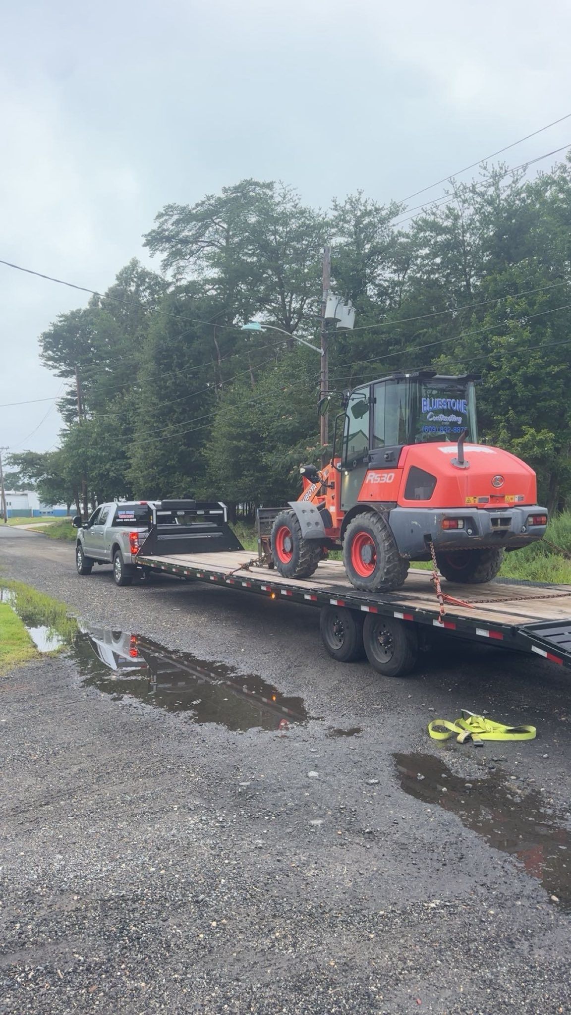 A silver pickup truck pulls a flatbed trailer loaded with a bright orange piece of construction machinery on a gravel lot.