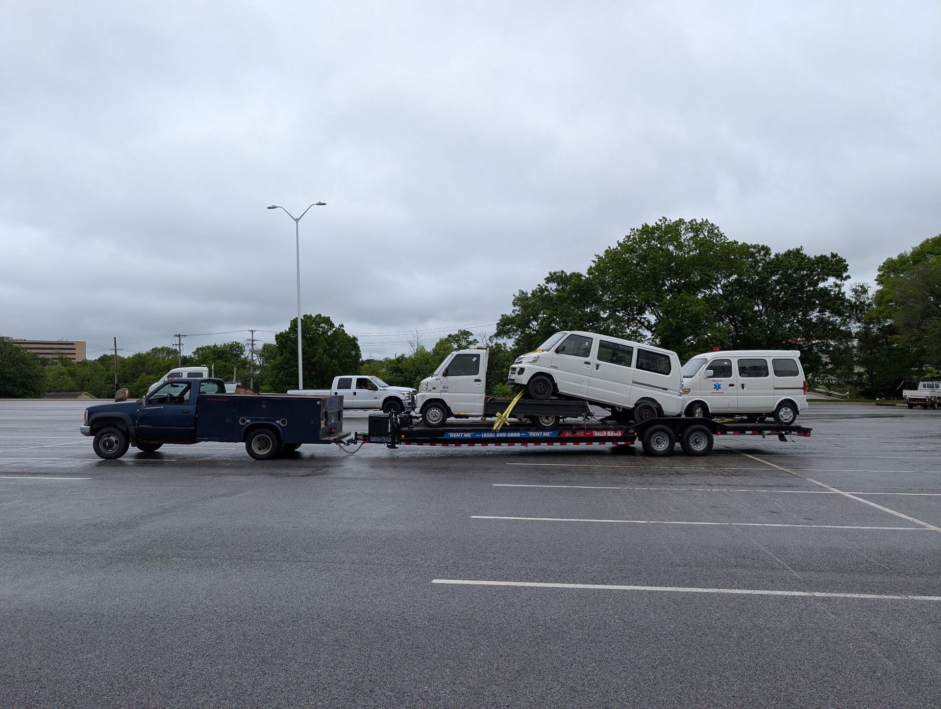 A blue pickup truck tows a car hauler carrying three white Kei vans across a large, wet asphalt parking lot.