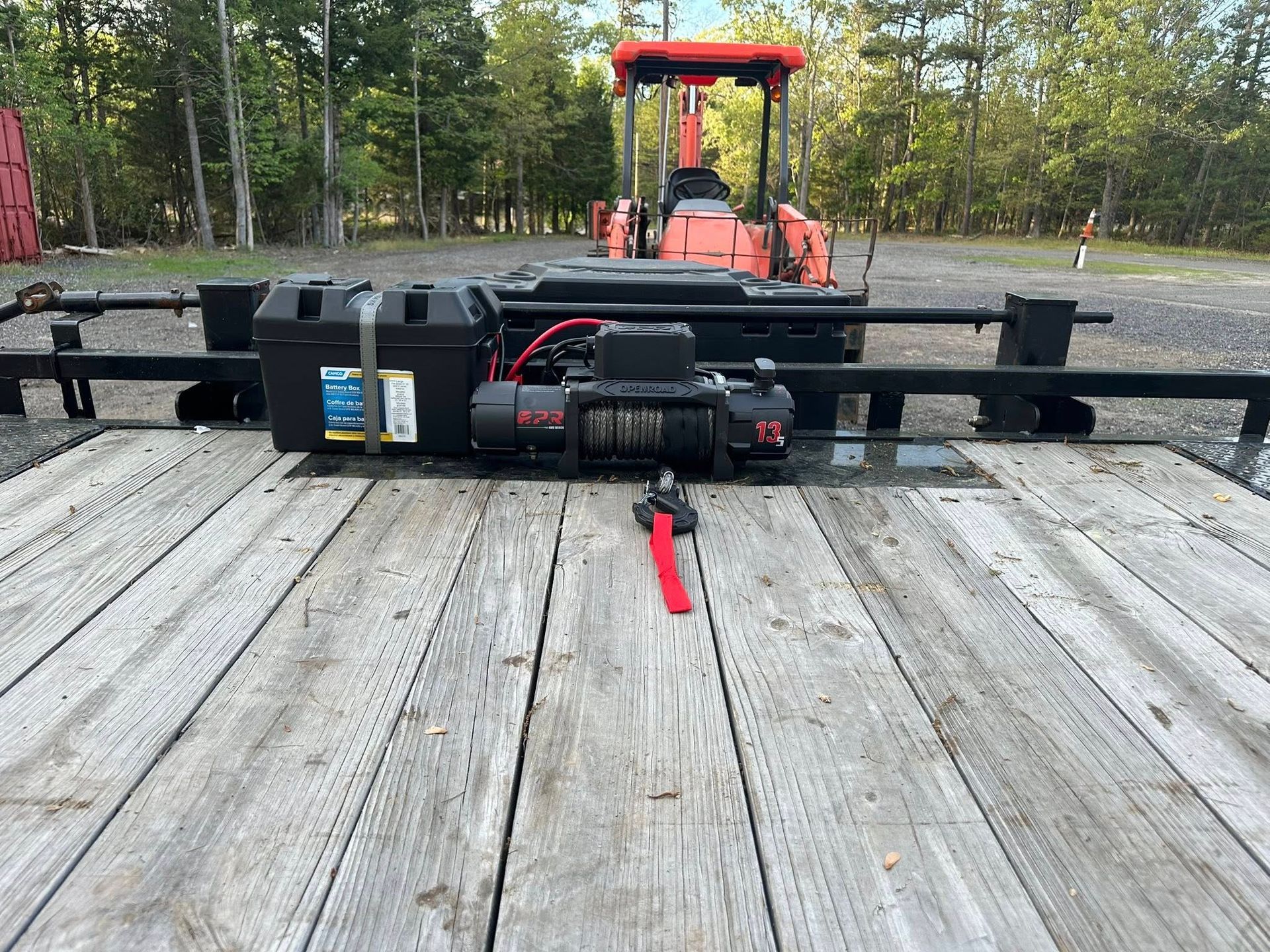 A winch and battery case mounted on the deck of a flatbed trailer, with an orange piece of machinery in the background.