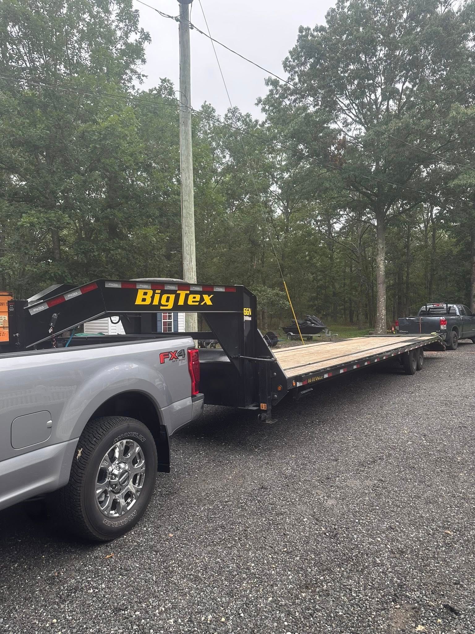 A silver pickup truck towing a black Big Tex gooseneck flatbed trailer parked on a gravel lot near trees.