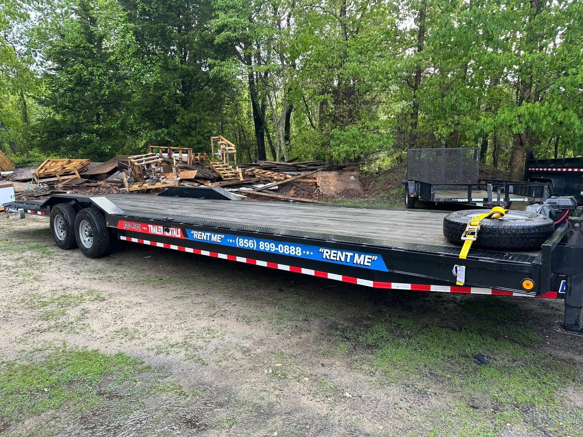 A black flatbed trailer parked on a gravel lot in front of trees, with a spare tire strapped to the rear deck.