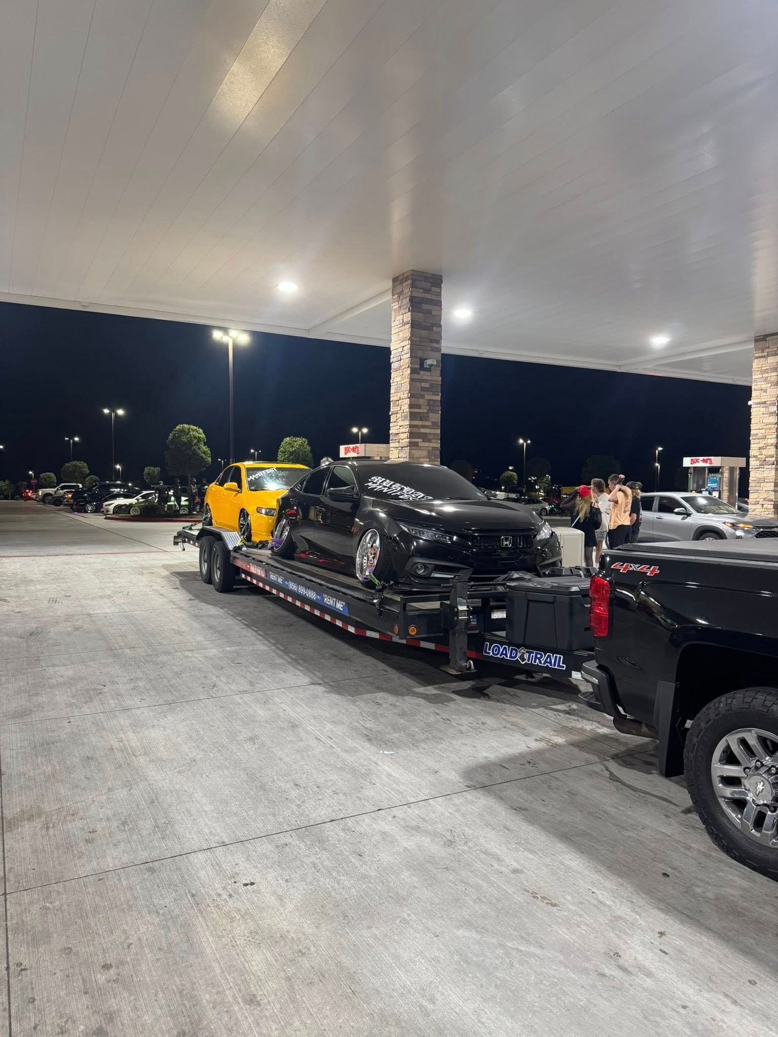 A black truck tows a trailer carrying a black car and a yellow car at a gas station at night.