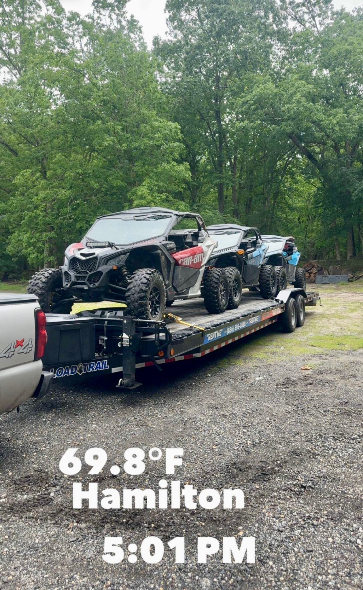 Two UTVs loaded on a flatbed trailer attached to a pickup truck parked on a gravel lot near a wooded area.