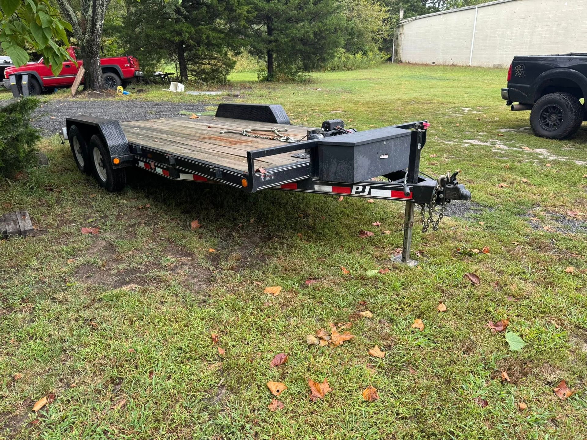 A dark metal car hauler trailer parked on a grassy lot, featuring a front storage box, jack stand, and red trim.