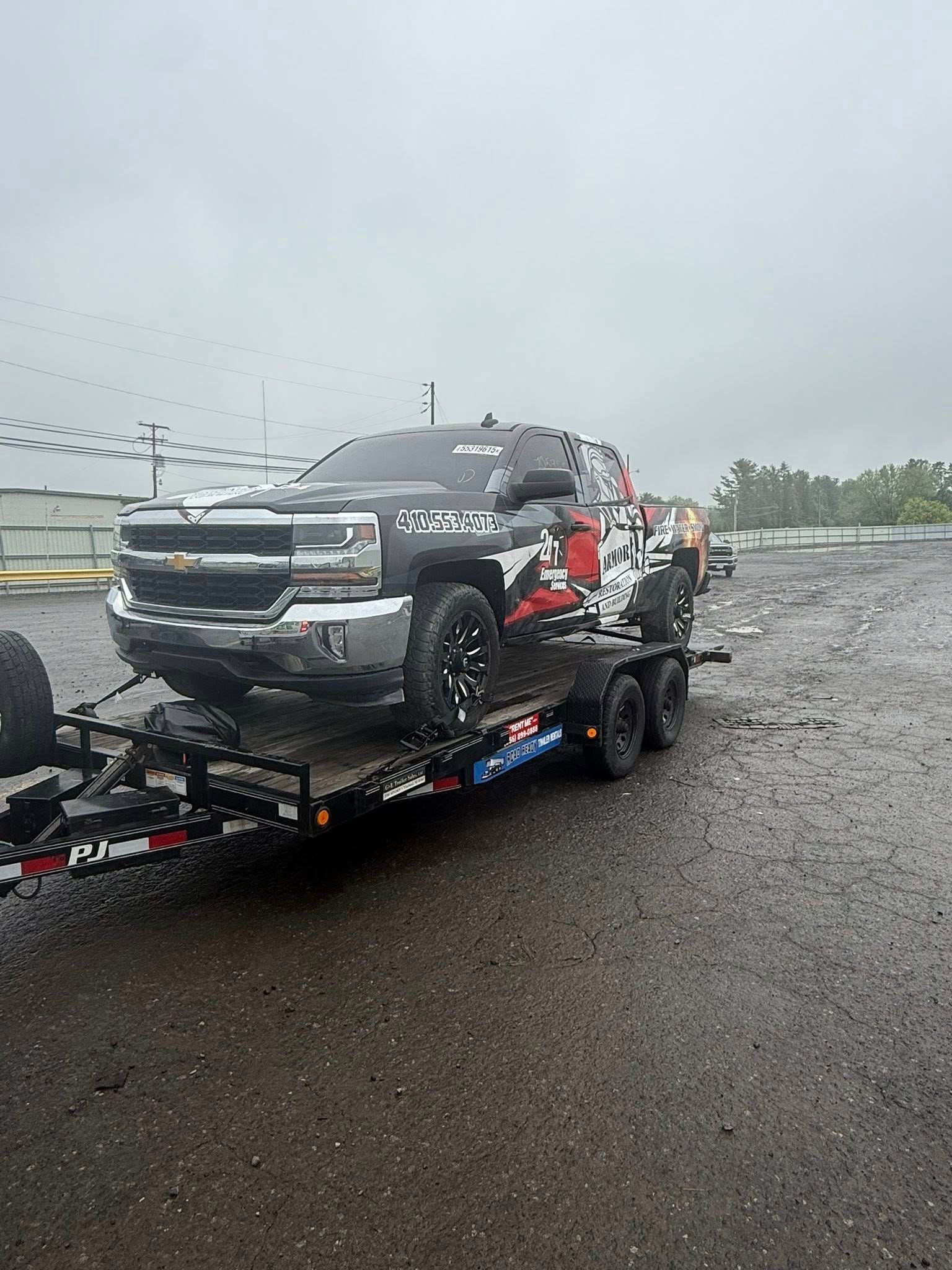 A black Chevrolet truck with a red and white camouflage pattern on a flatbed trailer on a gravel lot during rain.