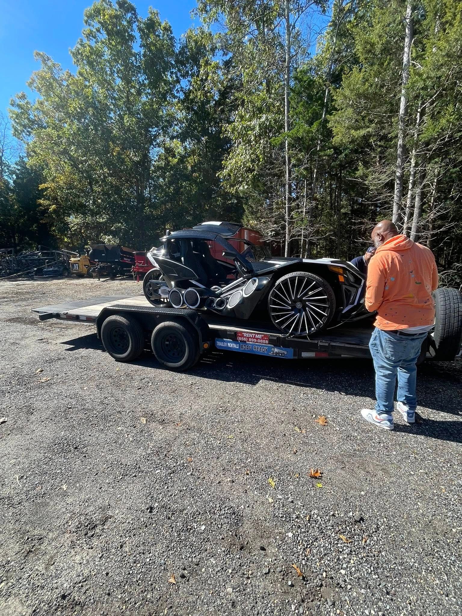 A person in an orange hoodie stands by a black three-wheeled vehicle strapped to a trailer in a gravel lot.