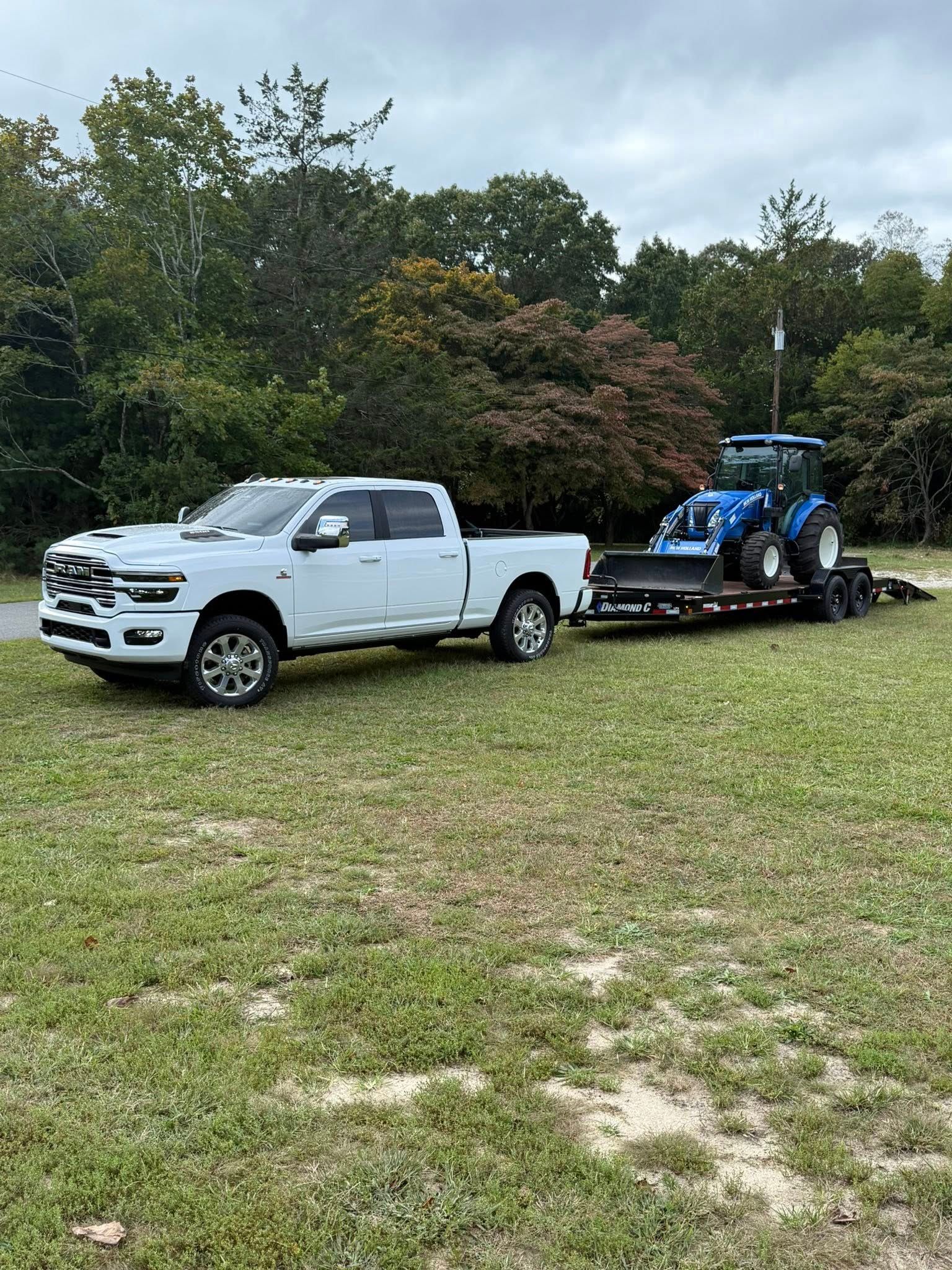 A white pickup truck towing a flatbed trailer with a blue tractor on it, parked on a grassy field with trees in the back.