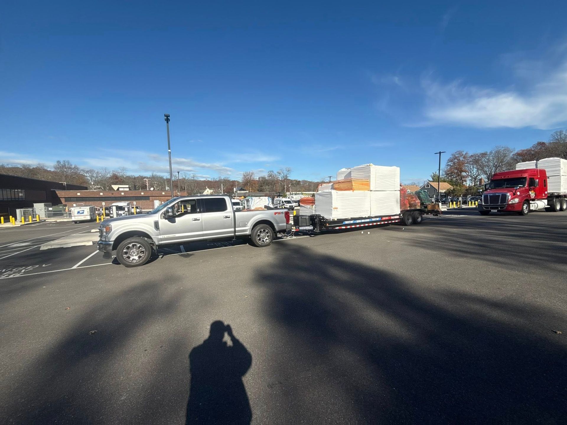 A silver pickup truck towing a flatbed trailer loaded with large, white, shrink-wrapped pallets in a parking lot.