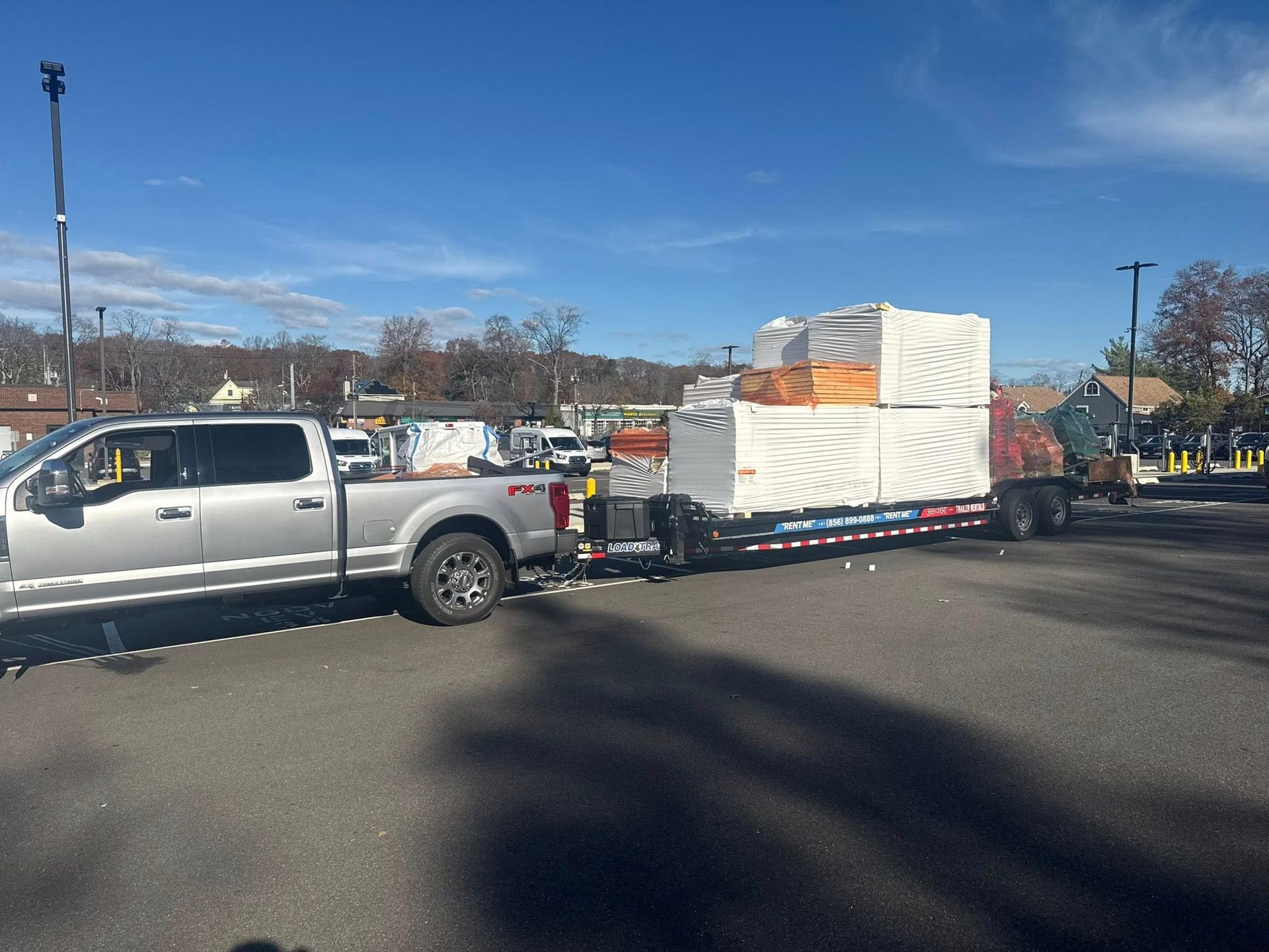 A silver pickup truck parked in an outdoor lot, towing a flatbed trailer loaded with large, white-wrapped palletized goods.