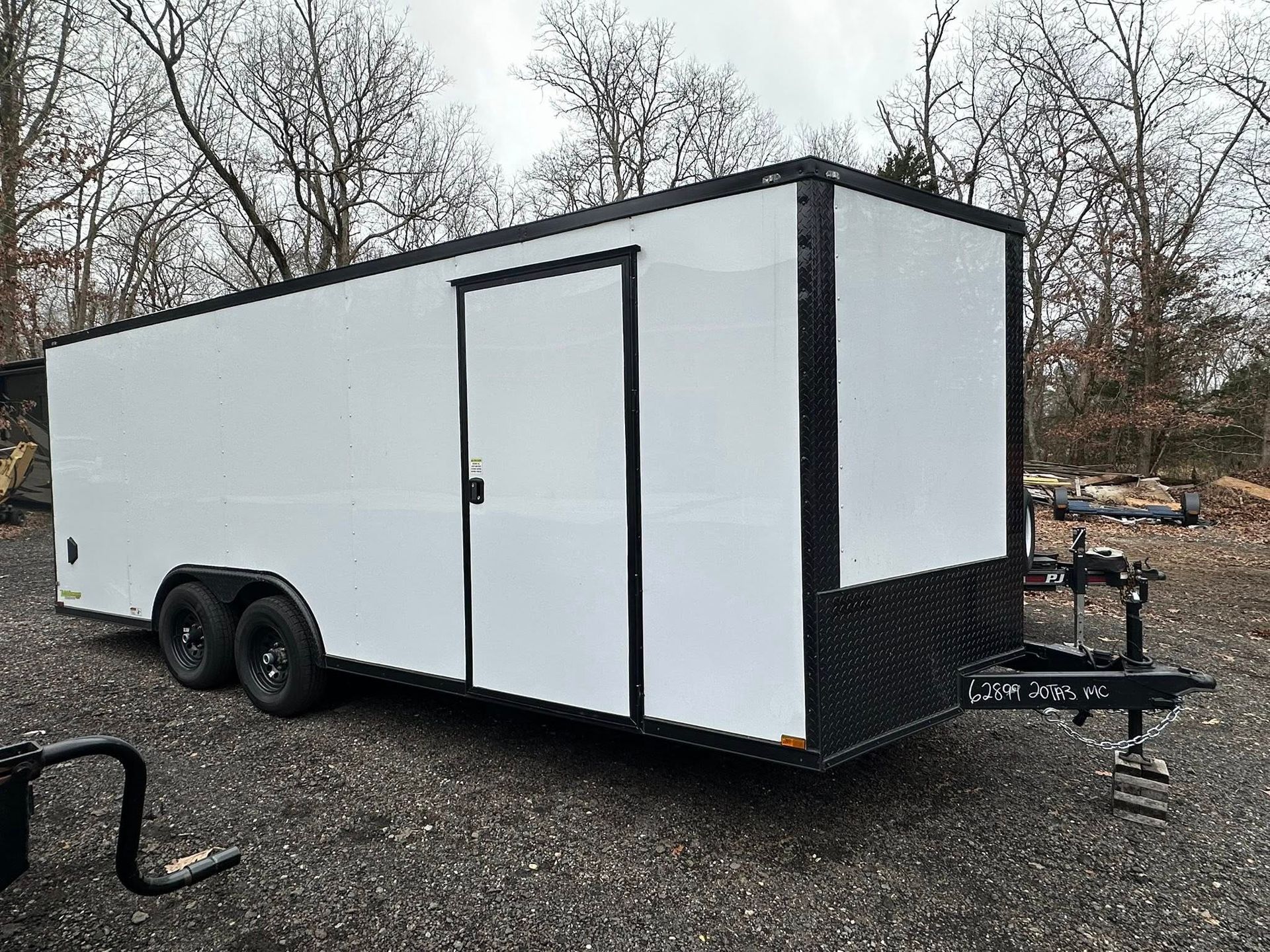 A white enclosed cargo trailer with black trim and wheels sits on a gravel lot against a backdrop of bare trees.