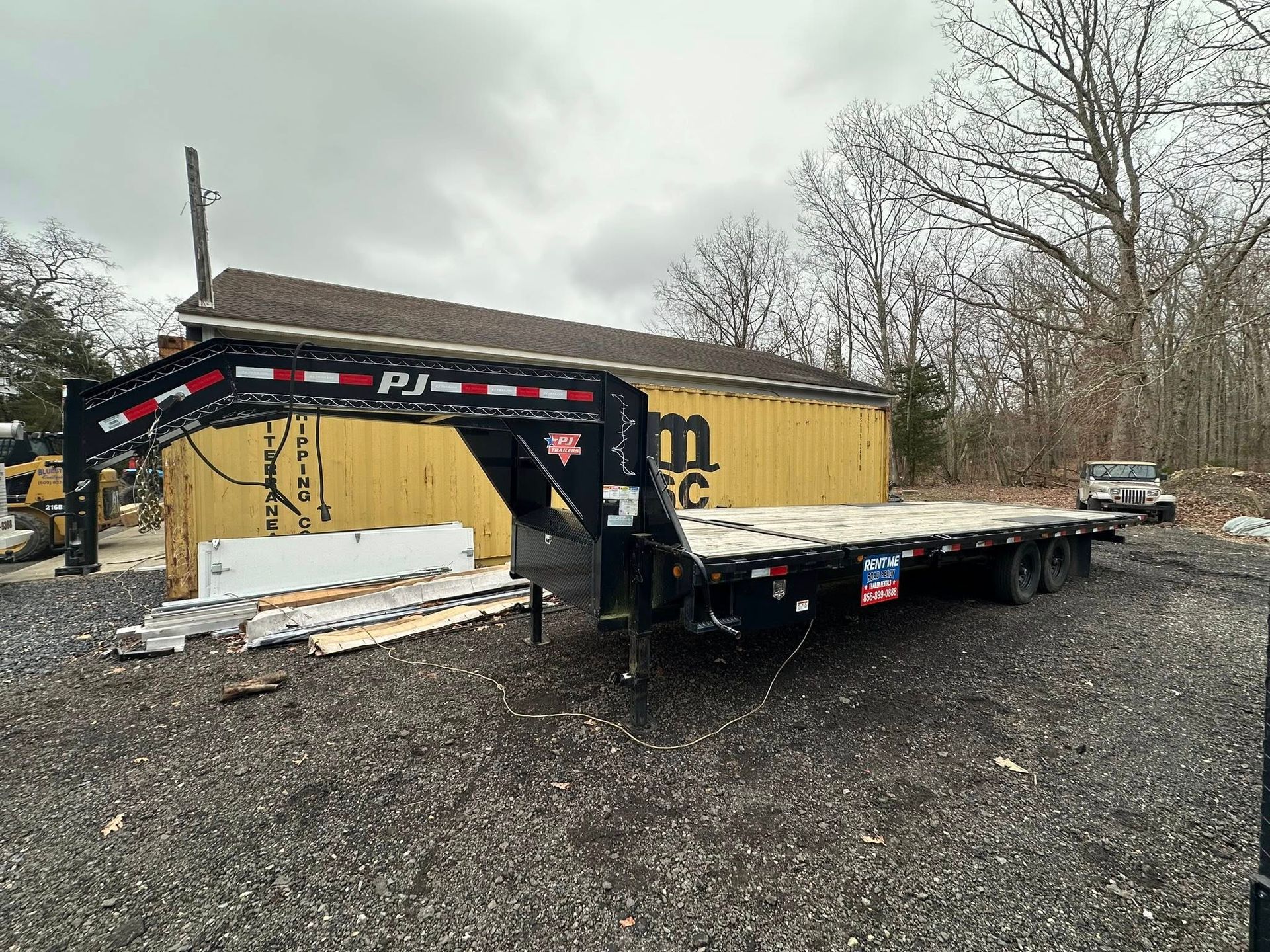 A black PJ gooseneck flatbed trailer parked on a gravel lot in front of a yellow shipping container.