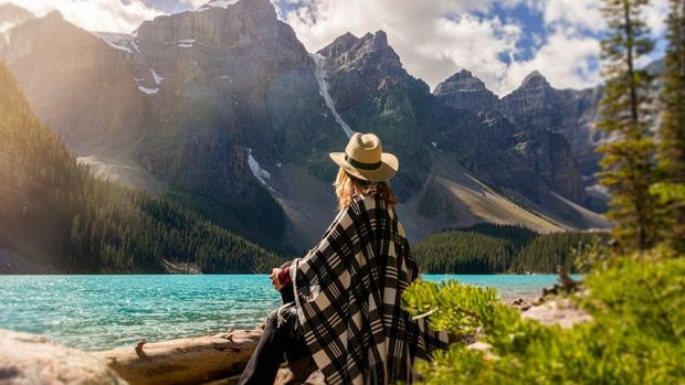 A woman is standing next to a lake with mountains in the background.