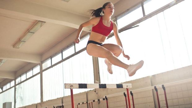 A woman is jumping over a hurdle in a gym.