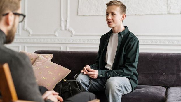 A young man is sitting on a couch talking to a man.