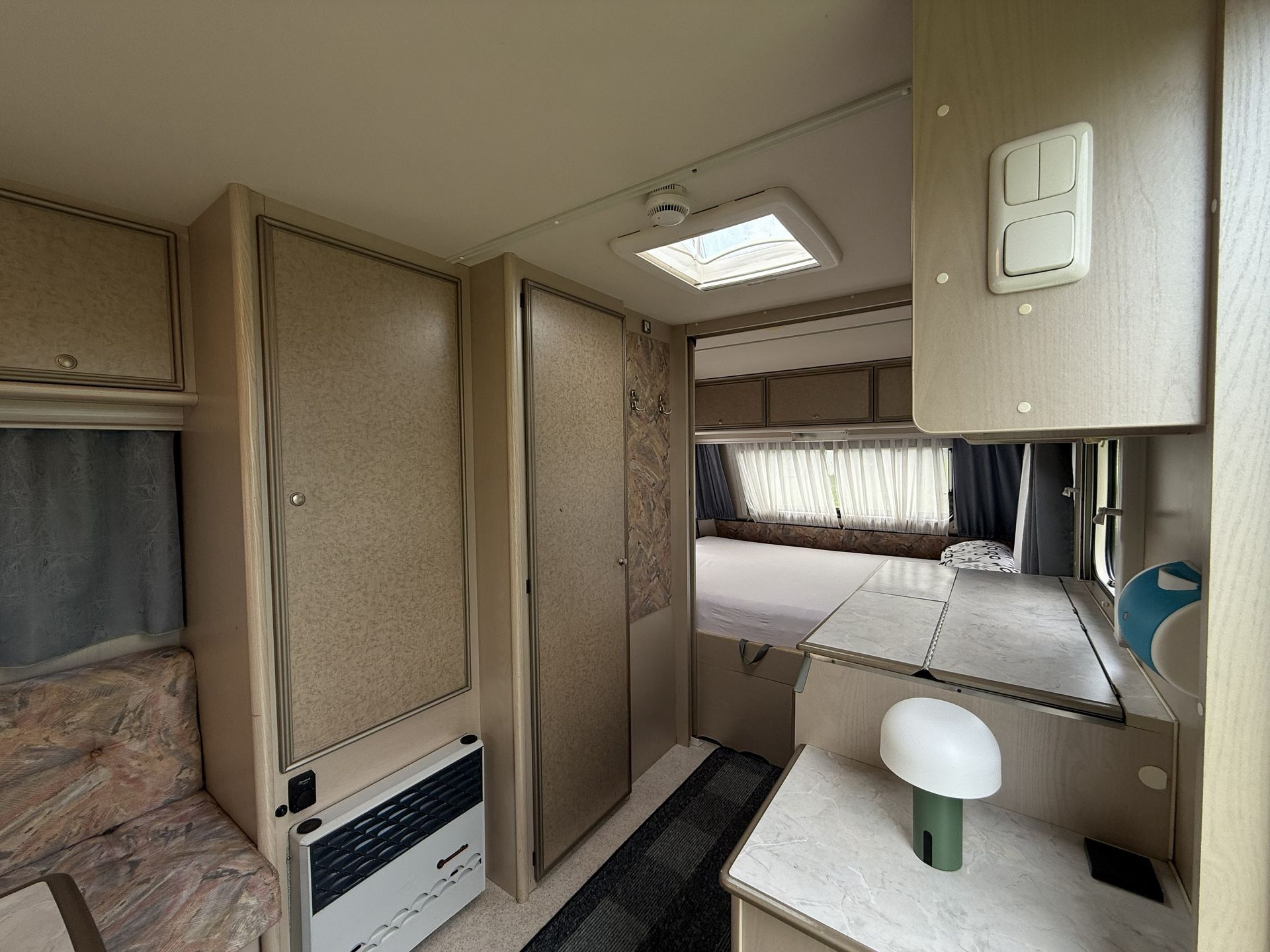 Interieur eines in neutralen Tönen gehaltenen Wohnwagens mit Bett, kleiner Küchenzeile, Stauschränken und einer Lampe auf einer Ablage.