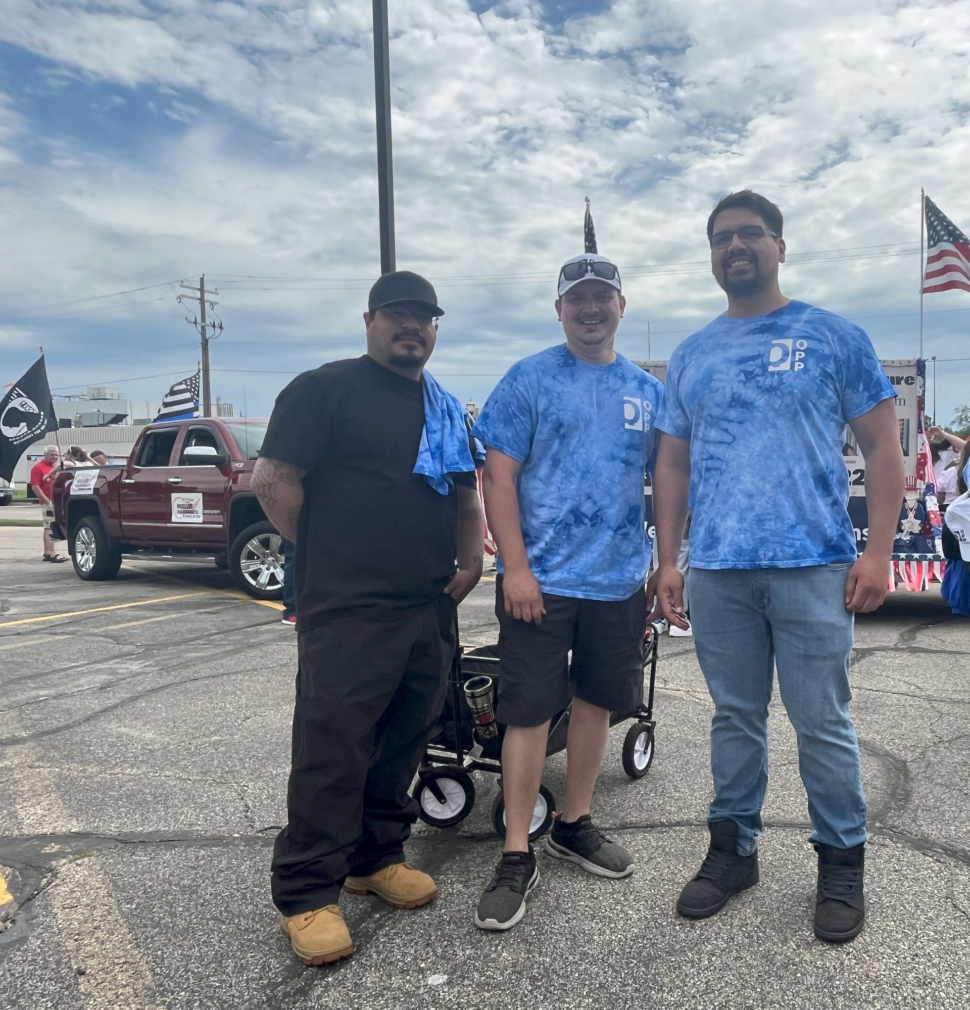 Three people pose outdoors in a parking lot, with a pickup truck and American flags visible in the background.