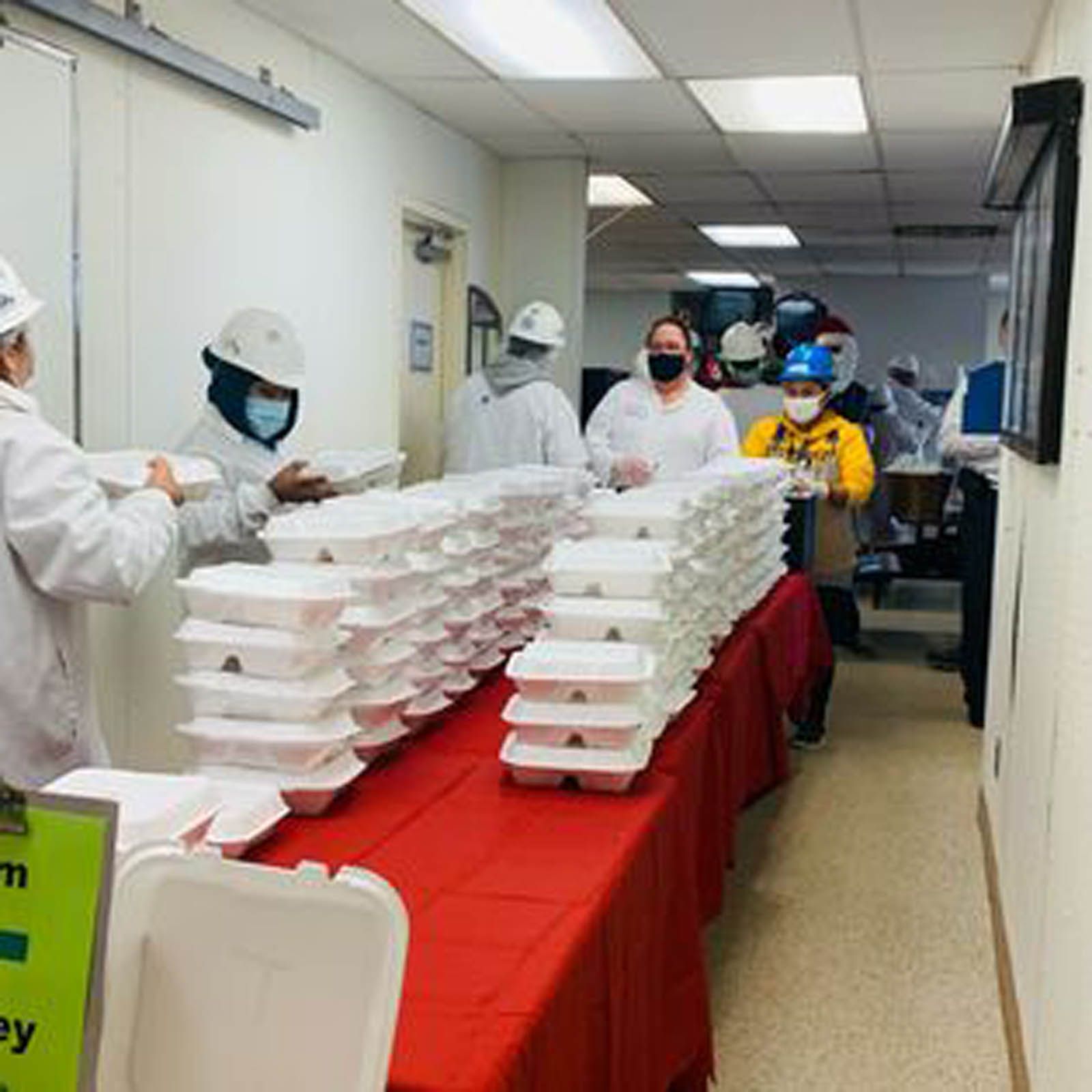 People in safety gear and face masks assemble stacks of white food containers on a red-covered table in a hallway.