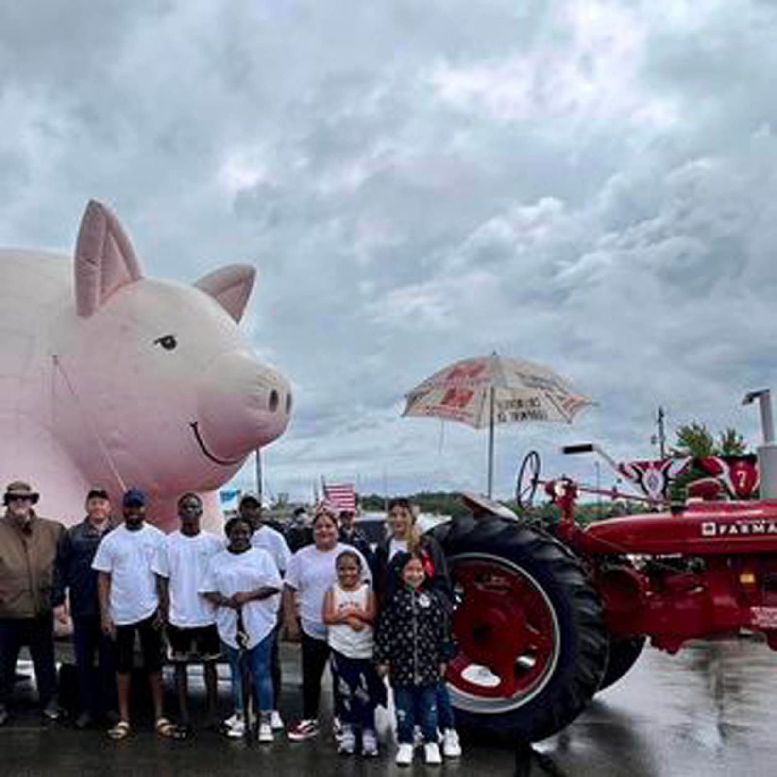A group poses outdoors with a giant inflatable pig and a red Farmall tractor under an overcast sky.