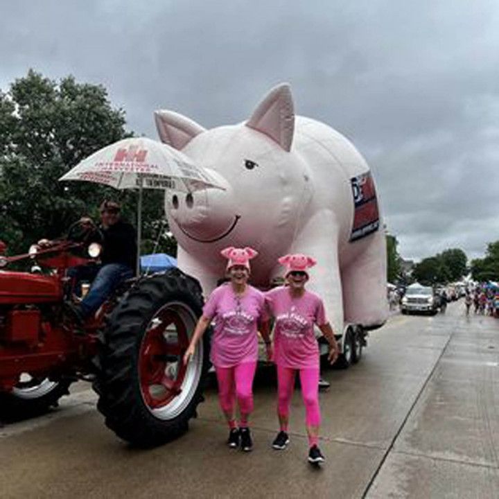 A red tractor pulling a giant inflatable pig, followed by two people in matching pink shirts and pig-ear headbands.