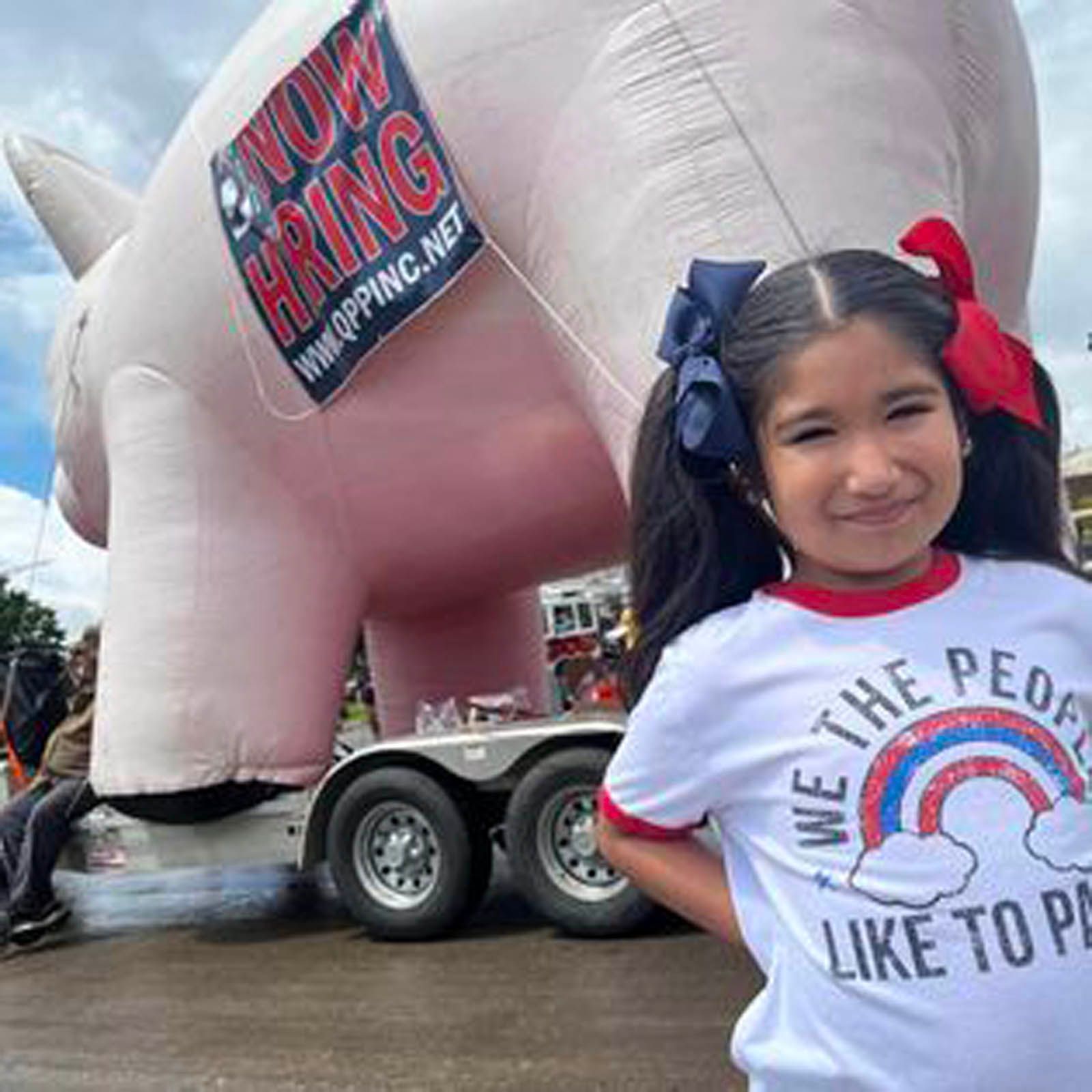 A smiling child in a red, white, and blue shirt stands in front of a giant inflatable pig with a 