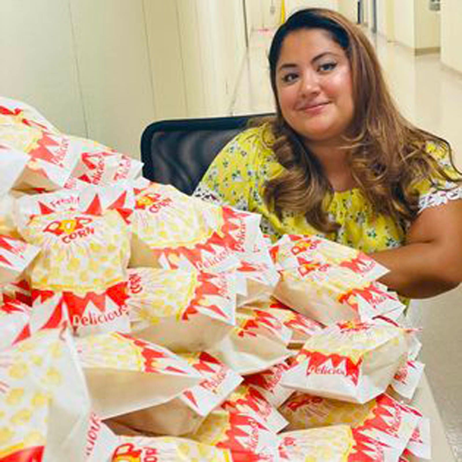 A person in a yellow floral top smiles behind a table piled high with numerous individual bags of popcorn.