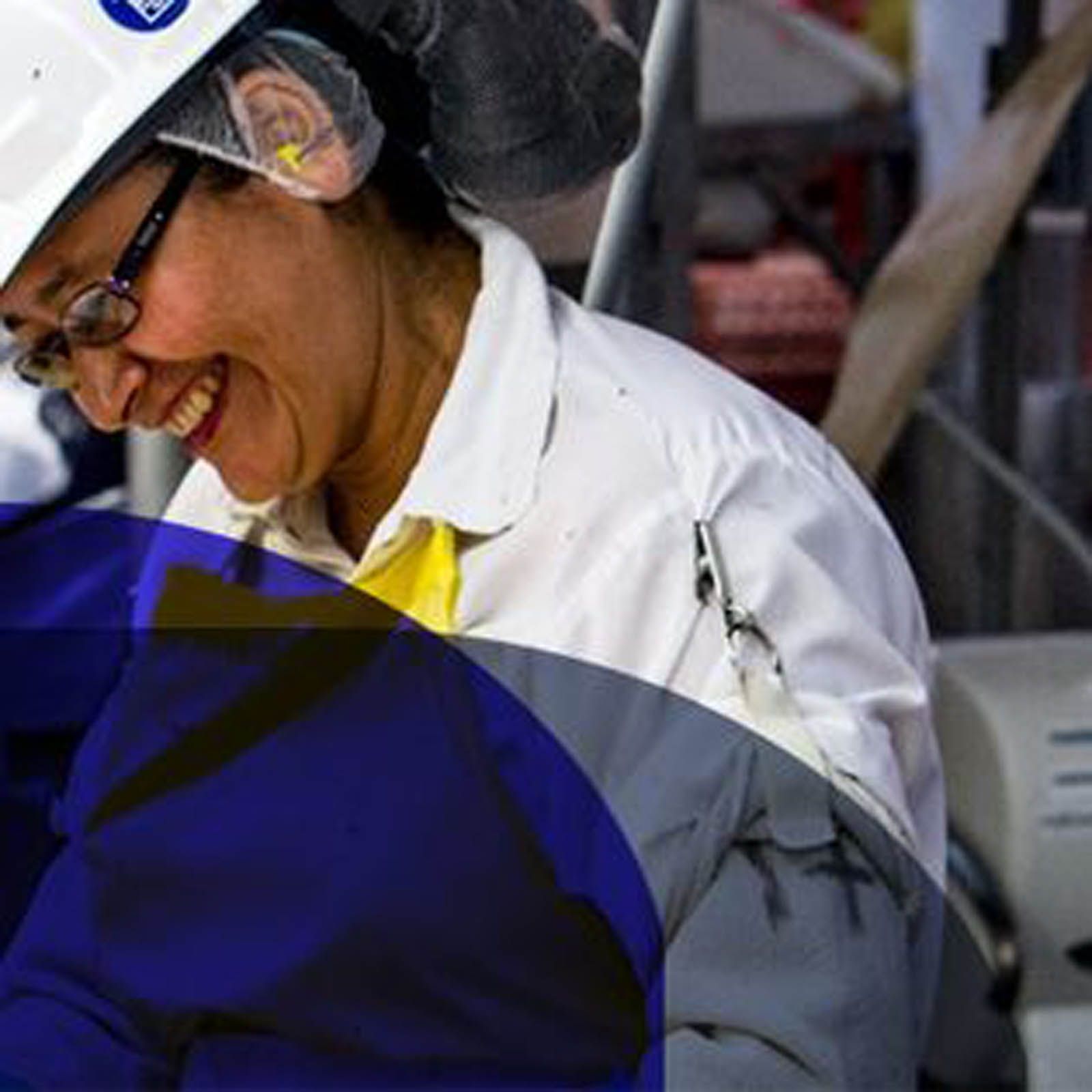 A person wearing a white hard hat and safety glasses smiles while working in an industrial setting.