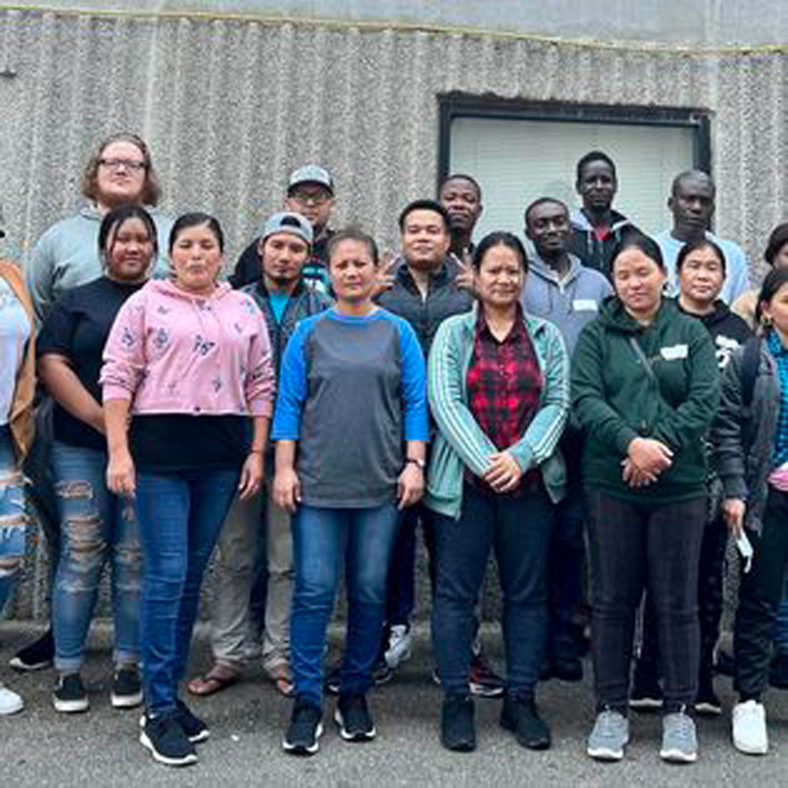 A group of seventeen diverse people stand in rows for a photo against a grey, textured wall and a rectangular window.