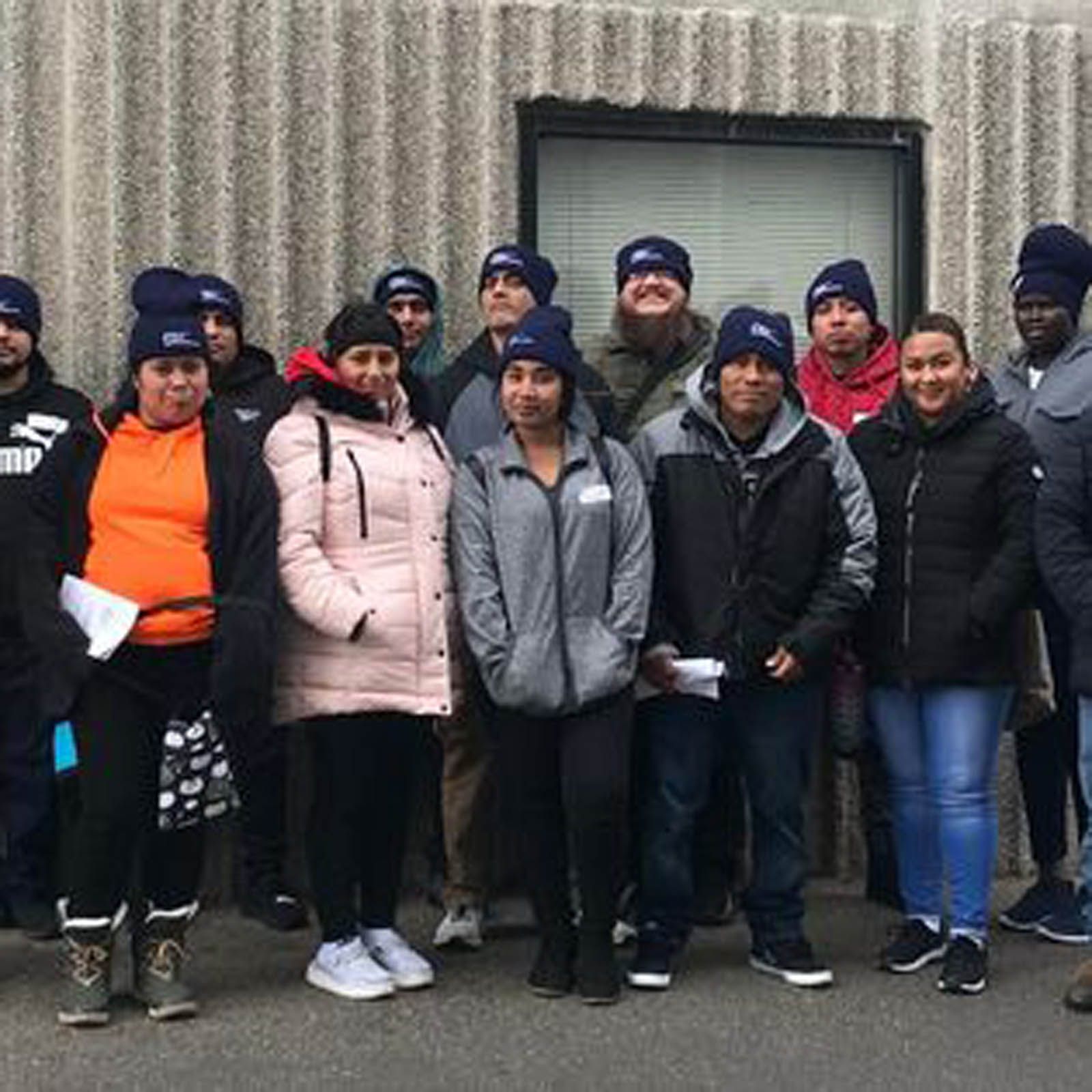 A group of people wearing matching navy beanies stands outside against a corrugated wall.