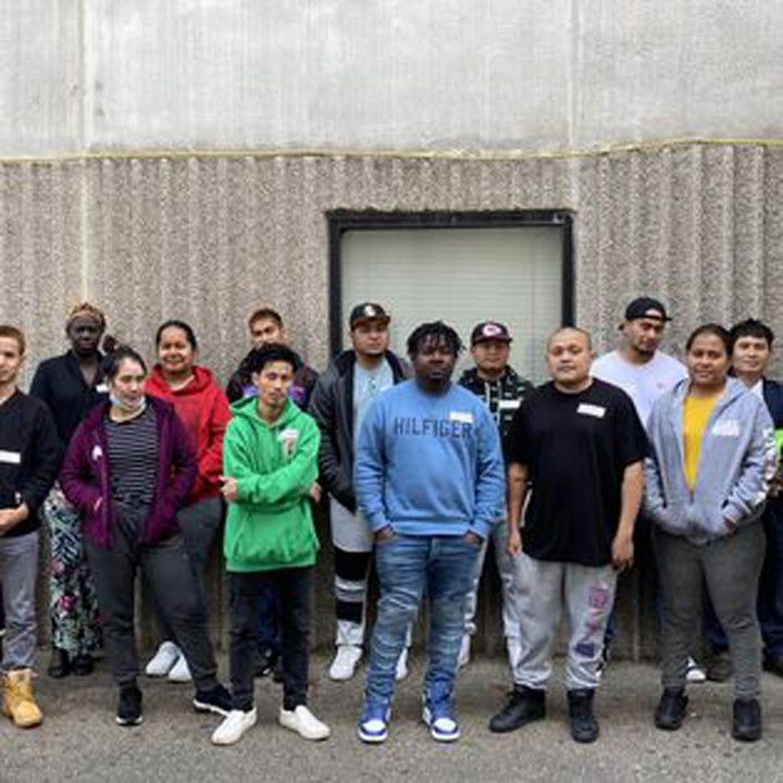 A group of fifteen people stand in front of a textured gray wall, posing for a photo outdoors.