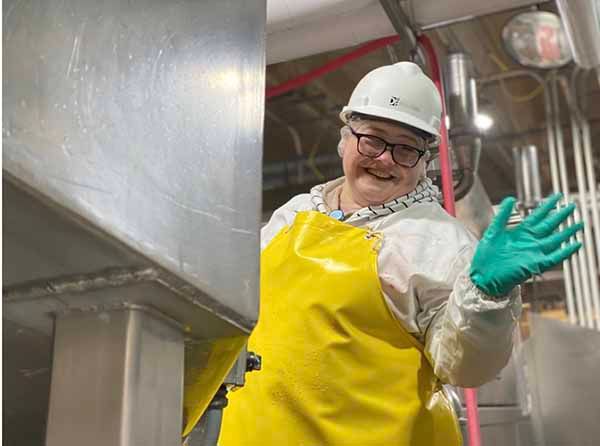 A smiling person in a hard hat, white coat, and yellow apron waves while standing in an industrial facility.