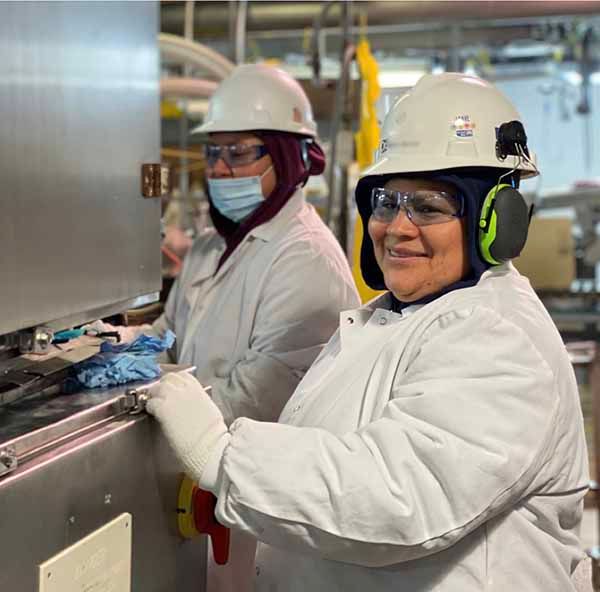 Two workers in white lab coats, protective helmets, and safety glasses operate industrial machinery in a plant.