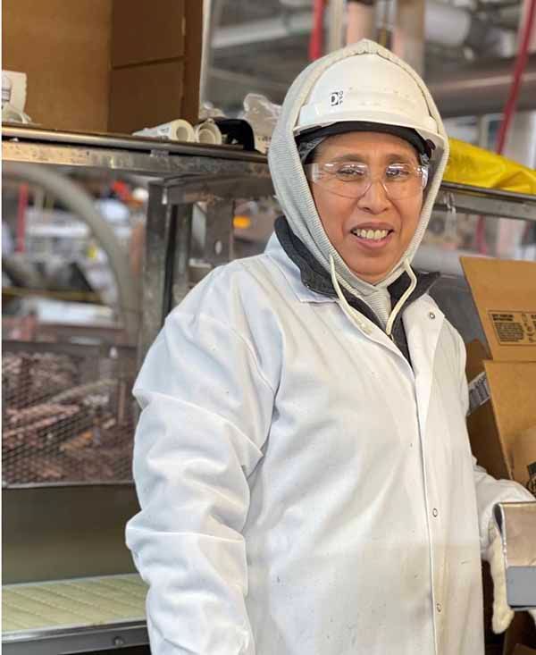 A worker in a white coat, hood, hard hat, and safety glasses smiling in a warehouse setting.