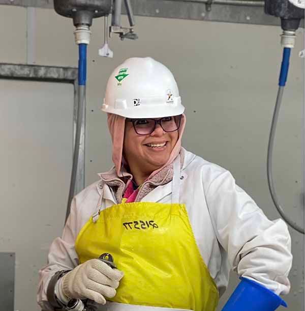 A smiling person in a white hard hat, face mask, and yellow apron holding a tool in an industrial setting.