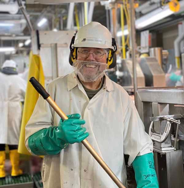 Smiling worker in a white lab coat, hard hat, safety glasses, and green rubber gloves holds a cleaning tool in a facility.