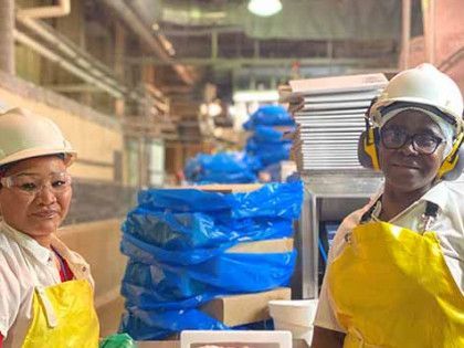 Two workers in white uniforms, yellow aprons, and hard hats stand in a brightly lit industrial food processing facility.