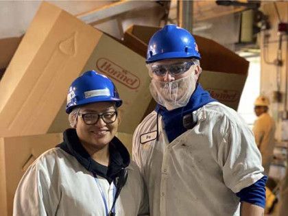 Two people in blue hard hats, safety glasses, and white smocks smiling in a warehouse with Hormel boxes in the background.