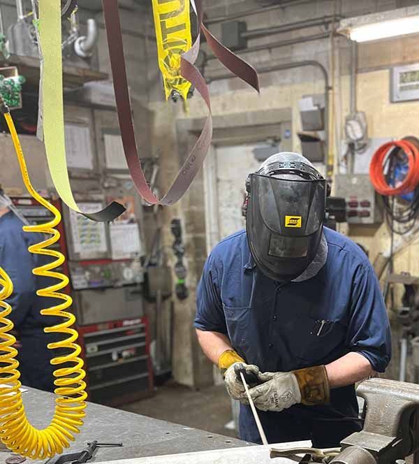 A person in a welding mask and safety gear works on a piece of metal in a workshop, with a yellow coiled air hose nearby.