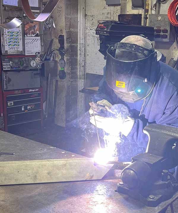 A person in a welding helmet uses an electric welder to join metal in a workshop, emitting a bright glow.