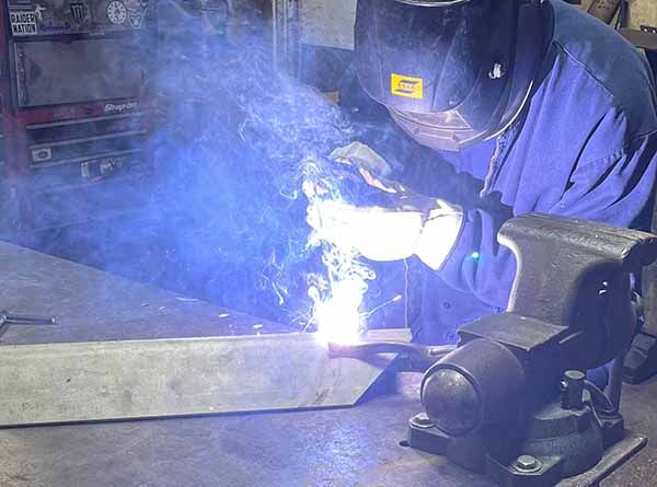 A person wearing a welding helmet and protective gloves uses a welding torch on metal near a bench vise.